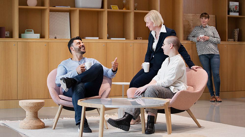 The work team - a man, a woman and non-binary person - are sitting together in the lobby of the workplace. A female colleague is smiling in the background.