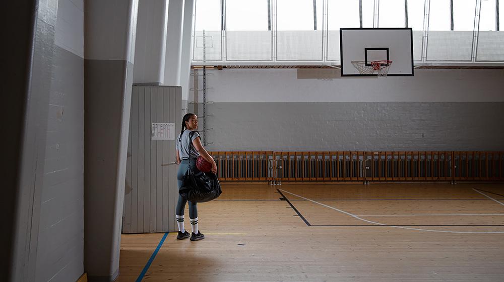 A young girl stands alone in an empty gym with a basketball under her arm.