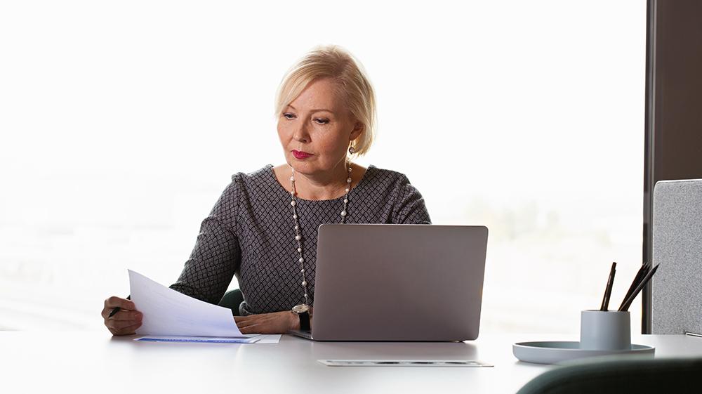 Nainen tietokoneen äärellä lukee papereita. 